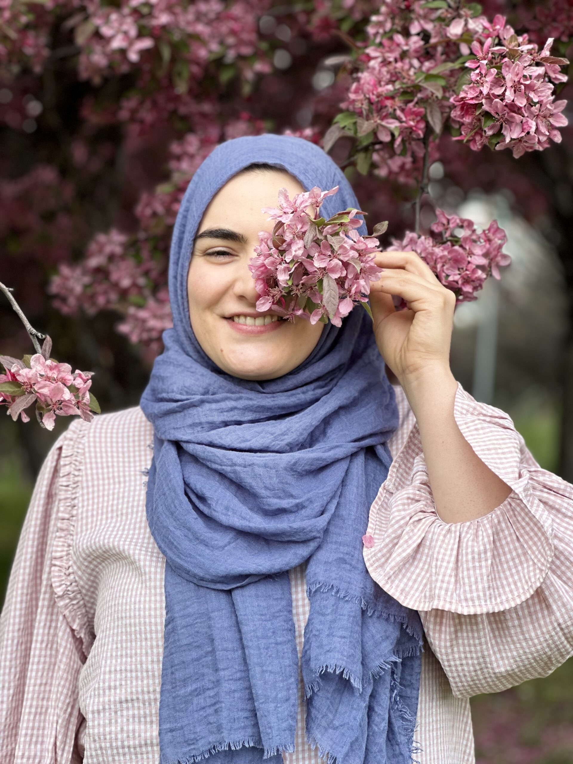 A woman wearing a light blue headscarf and a light pink ruffled blouse stands in front of a tree full of pink blossoms, gently holding a cluster of flowers in front of one eye while smiling | Germanised Gülnur